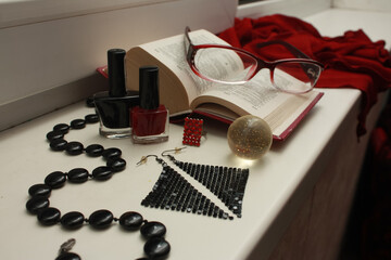 Composition of red glasses, black beads made of natural agate, nail polishes, shiny disco earrings, a transparent shiny ball, an open dictionary, a matrix ring, on a white windowsill.