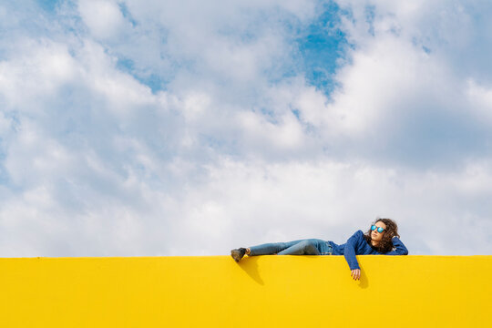Young woman relaxing on yellow wall against cloudy sky