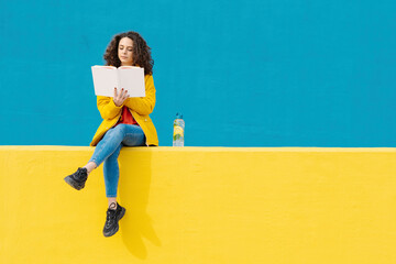 Young woman sitting on yellow wall reading a book