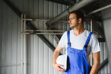 Smiling and happy employee. Industrial worker indoors in factory. Young technician with white hard hat.