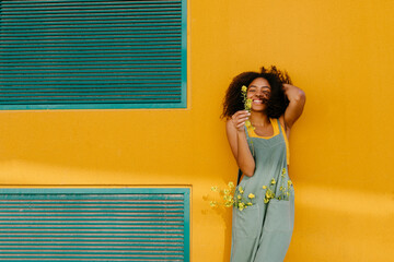 Portrait of happy young woman wearing overalls holding flowers in front of yellow wall