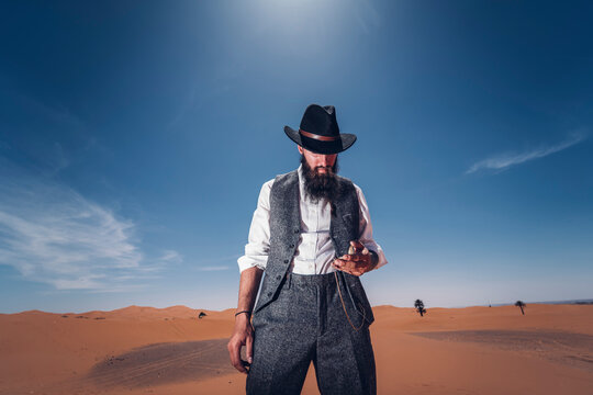 Man with a beard and hat in the dunes of the desert of Morocco checking the time