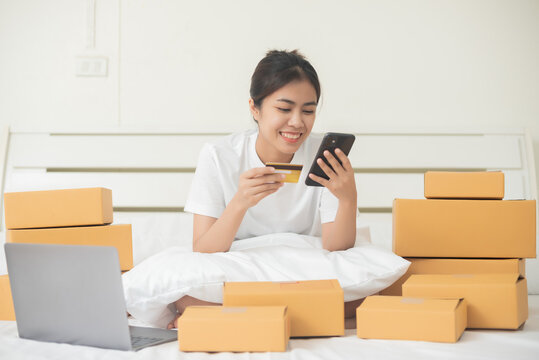 An Asian woman happily pays with a credit card on her smartphone in her bed at home. Around her, there are a lot of boxes ordered from the online website.