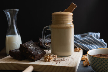 Dalgona coffee cup with a cookie on top on a wooden board with brownies and nuts, a jug of milk and...