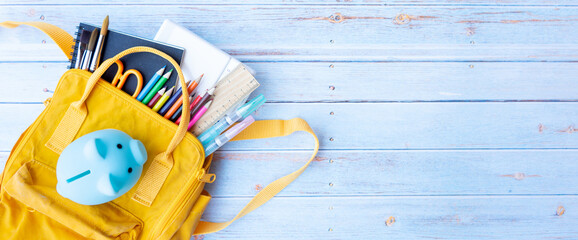 Top view backpack with different colorful stationery and piggy bank on the wooden table with copy space. Back to school concept