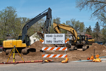 Road Closed sign with heavy construction equipment in the background framed by trees and a blue sky.