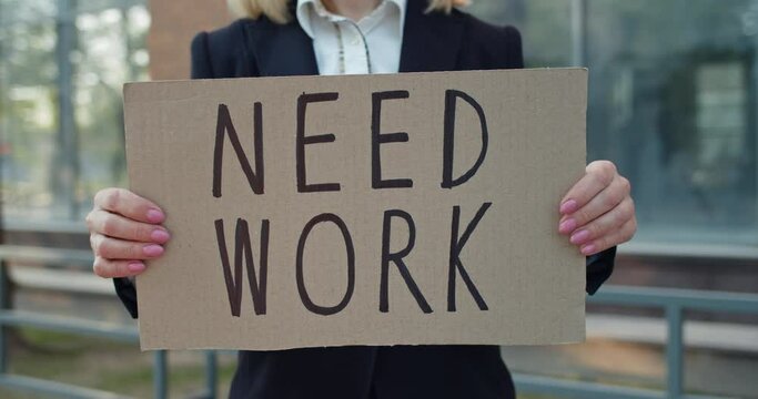 Crop view of female worker showing carton banner with need work writing. Woman searching for job while standing at street. Concept of financial crisis and unemployment