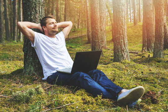 young man with laptop leaning against the tree and relaxing in the park