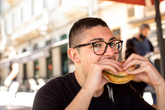 ragazzo con occhiali mangia un panino con cotoletta seduto al tavolino di un bar
