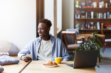 Millennial African American guy with smartphone and laptop enjoying morning coffee and croissant in cafe