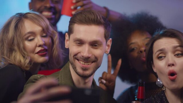  Slowmo handheld shot of happy young man holding mobile phone and taking selfie with multiethnic group of friends at party