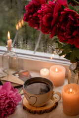 Cup with coffee on a windowsill with candles and flowers. Rain outside the window.