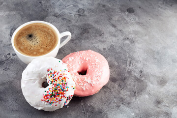 White cup of coffee and two donuts in white and pink icing and multicolored sugar pastry topping on grey background. Copy space