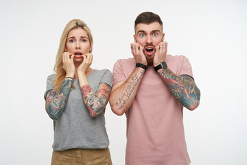 Shocked young pretty tattooed couple holding raised hands on their cheeks while looking amazedly at...