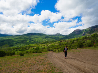 Obraz premium A young girl in a red tank top with a gray backpack in the mountains with green trees and clouds.