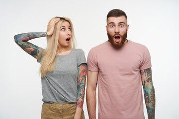 Shocked young attractive tattooed couple in casual t-shirts keeping their mouths wide opened while being amazed with something, standing against white background