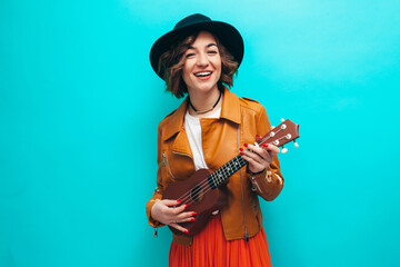 Handsome young hipster girl wearing black leather jacket and a hat plays ukulele guitar isolate...