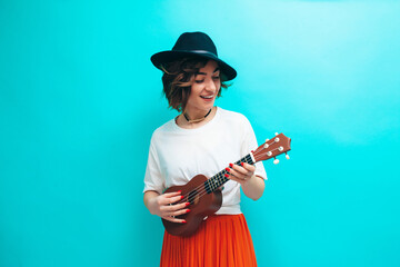 Obraz premium Handsome young hipster girl in casual clothes plays ukulele guitar isolate over blue background.