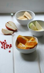 large pieces of different fruits in white bowls are located on a textured light wooden background