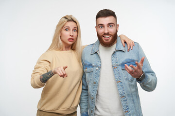 Fototapeta premium Agitated young pretty couple in casual wear rounding their blue eyes while looking amazedly at camera and raising surprisedly eyebrows, isolated over white background