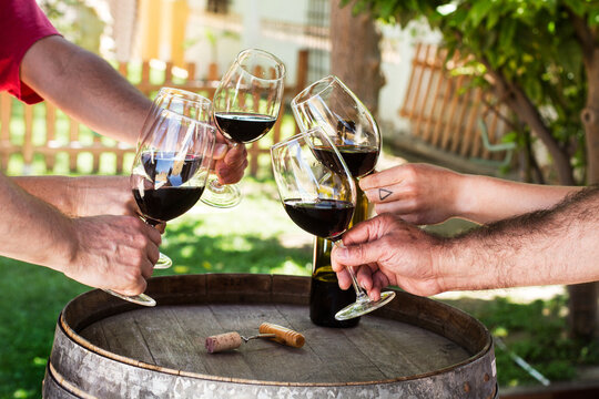 Un grupo de manos brindando con copas de vino tinto junto a un rústico barril en una terraza al aire libre. Vista de frente y de cerca