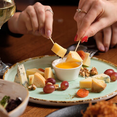 Female hand holding canape near cheese platter with different cheese, honey and grapes on a table for brunch. female friends - hands clinking white wine glasses,