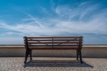 Naklejka na ściany i meble An empty bench against the background of blue sky and clouds in the Park stands on gray paving stones, in front of the parapet.