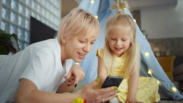 Pleasant Caucasian mother and daughter connected online in internet using smartphone, positive family parent and kid playing games in application holding mobile device leisure sharing content