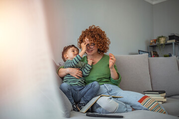 Working at home mom. Young modern mother with a toddler boy study at home. Mom studies for college exam with her boy. Young red haired woman working from home.