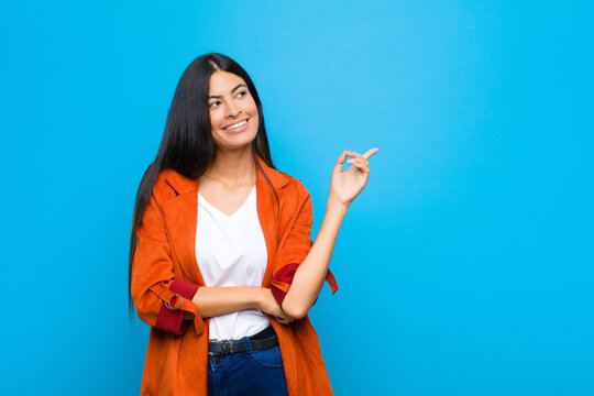 young pretty latin woman smiling happily and looking sideways, wondering, thinking or having an idea against flat wall