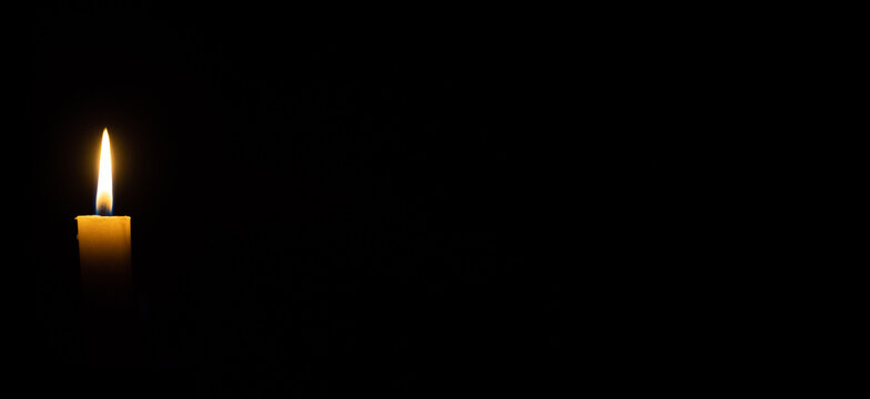 A candle burns in the darkness, copy space. A lit white candle on a black background. The symbol of the eternal memory, mourning, moment of silence, day of remembrance, of loss. Banner or site header