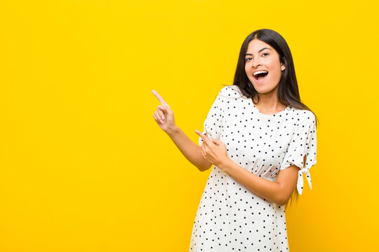 young pretty latin woman feeling joyful and surprised, smiling with a shocked expression and pointing to the side against flat wall