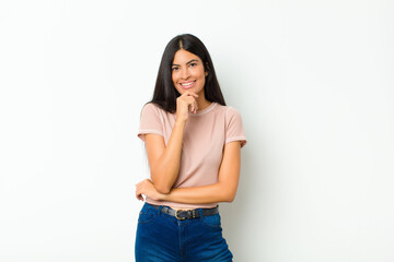 young pretty latin woman looking happy and smiling with hand on chin, wondering or asking a...