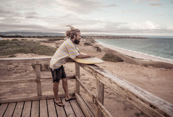 Portrait of Hipster Surfer with dreadlocks and beard looking at the ocean with vintage surfboard .