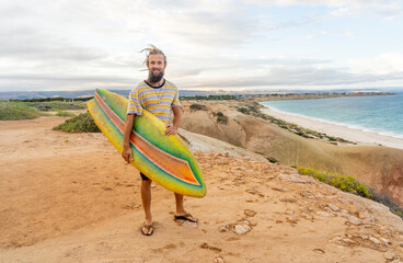 Portrait of Hipster Surfer with dreadlocks and beard looking at the ocean with vintage surfboard .