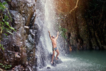 Happy young man relaxing under tropical waterfall with arms up raised in freedom. Health and relaxation.