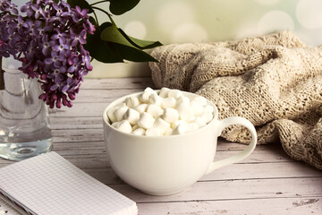 Marshmallows in a white mug, next to a vase with lilacs, is a notebook with place for text