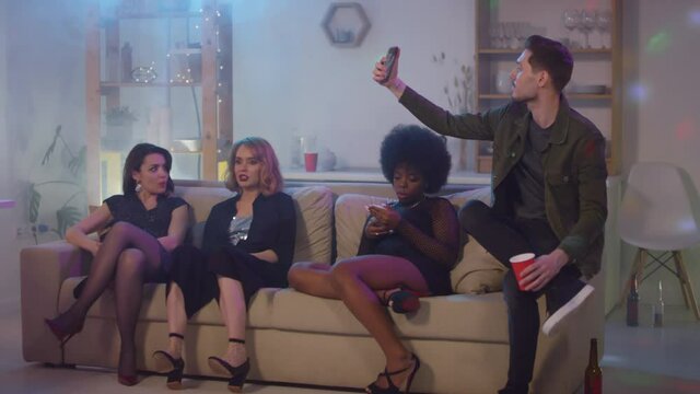  Slow motion dolly-in shot of young women sitting on couch at house party as bearded man taking selfie
