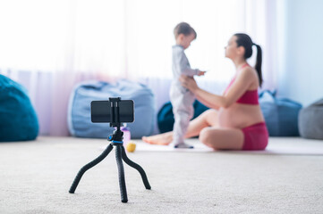Fototapeta na wymiar A pregnant woman does gymnastics with her son live. The expectant mother shoots a training video for her blog on the phone. Fitness with a baby.