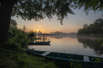 Beautiful early morning peaceful countryside landscape. Horizontal photography of old wooden rowboats in foreground, foggy water of small river, misty horizon line and dark trees growing at shores.