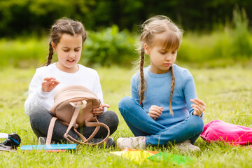 Fototapeta premium two cute multicultural schoolgirls sitting on lawn under tree and reading book together