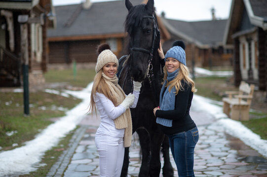 Picture of two young attractive sisters in warm jackets, trousers, boots, scarfs and knitted hats with a horse outside