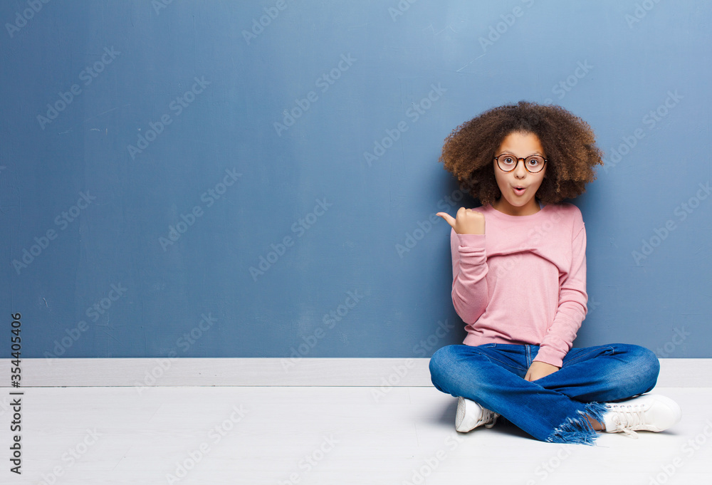 Wall mural african american little girl looking astonished in disbelief, pointing at object on the side and saying wow, unbelievable sitting on the floor