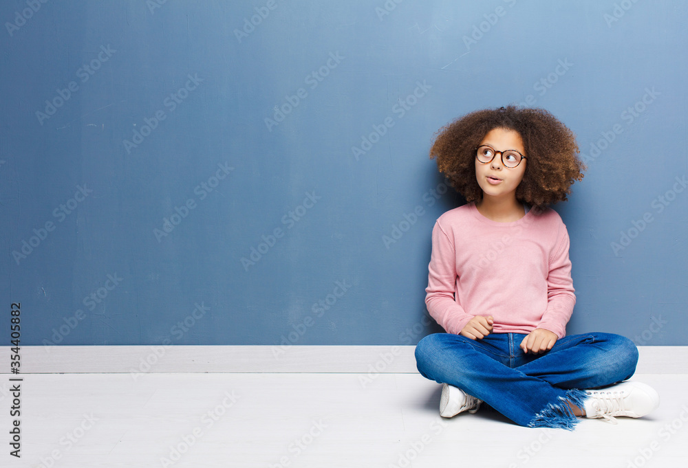 Wall mural african american little girl feeling shocked, happy, amazed and surprised, looking to the side with open mouth sitting on the floor