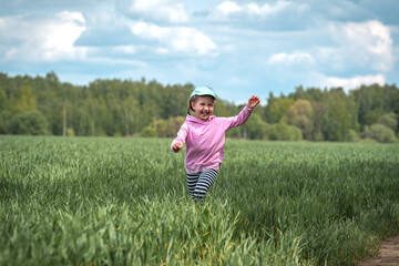 Fototapeta premium Funny little girl runs through field with green wheat shoots in countryside.