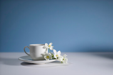 Obraz na płótnie Canvas White cup of morning espresso with a blossoming apple tree branch on a stone surface of a table with reflections opposite blue background.