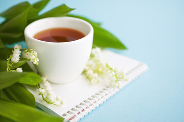 Cup of tea, notebook and a bouquet of lilies of the valley on a blue background, soft focus, copy space