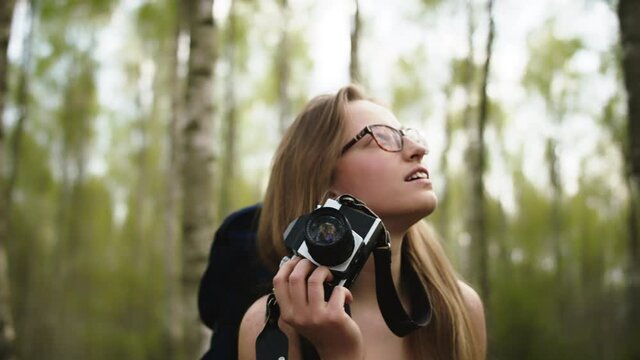 Happy young caucasian woman traveler with backpack and vintage camera surrounded by trees. Portrait slow motion shot.