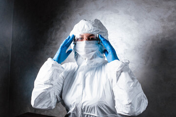 Virologist in white mask in a viral protective suit holds head, looks at the camera with fear and...