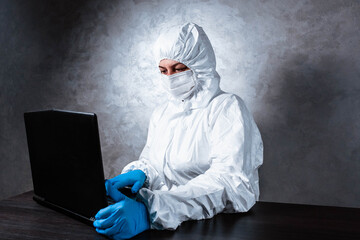 A virologist in a white mask and medical gloves dressed in a white protective suit works with a laptop and looks for information about coronavirus. Isolated and in panic due to epidemic.